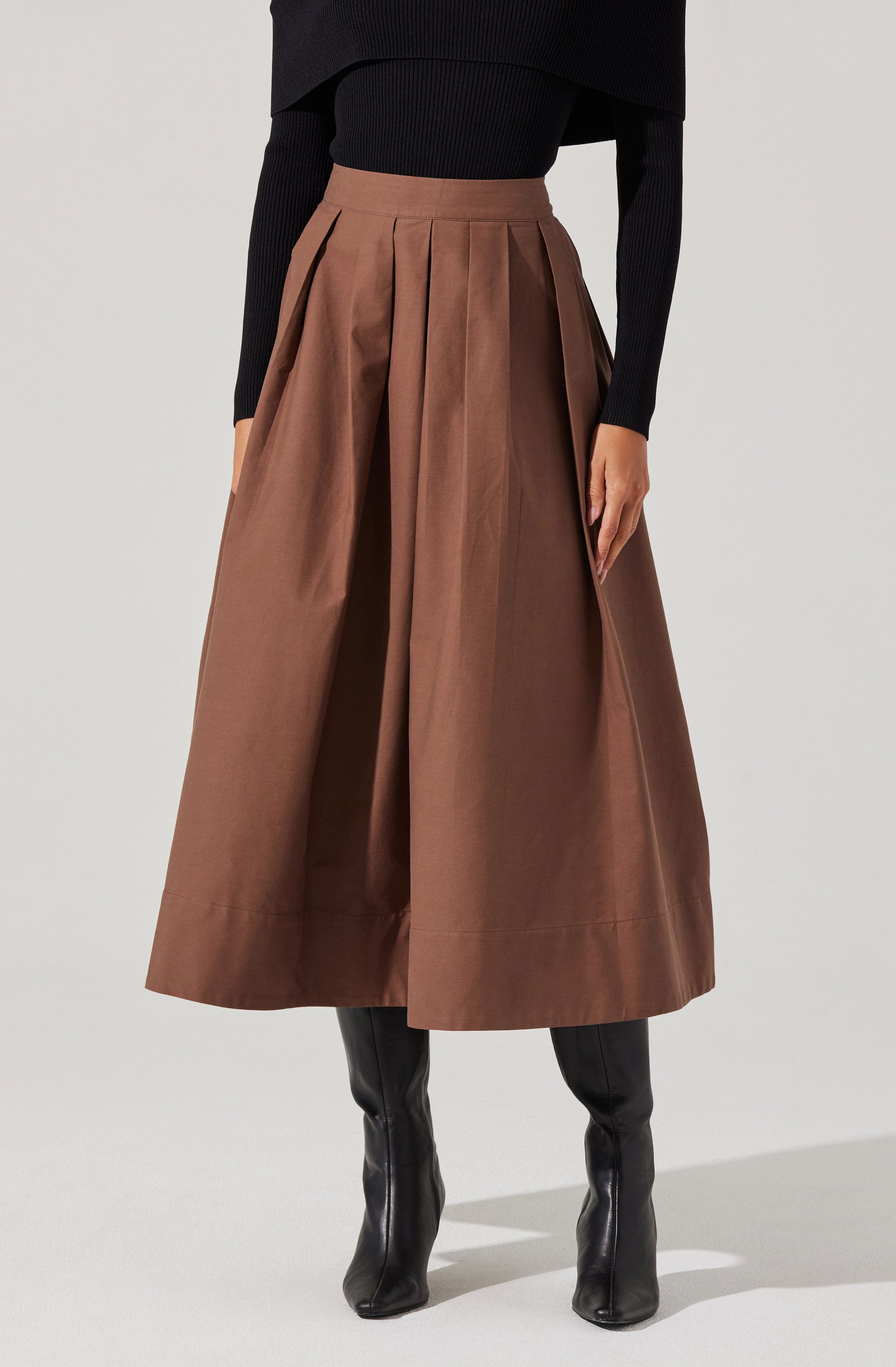 The outfit features a high-waisted, full-length brown skirt with pleats that add volume and structure. The fabric appears to be a sturdy, smooth material. The person is also wearing a fitted black long-sleeve top that is tucked into the skirt. The top has a ribbed texture and a slightly cropped style. The individual is wearing tall black leather boots that reach just below the knees.