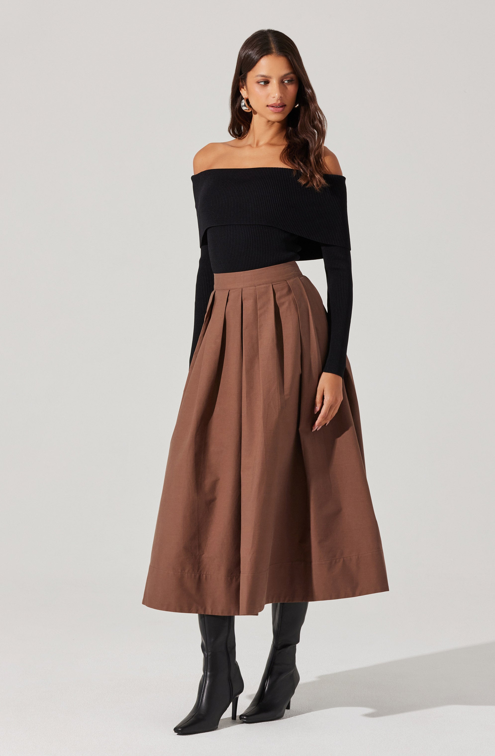 A woman is wearing a black off-the-shoulder top with long sleeves and a wide neckline, paired with a high-waisted, full-length brown skirt that features pleats and a slightly shiny, structured fabric. She completes the look with black heeled boots.