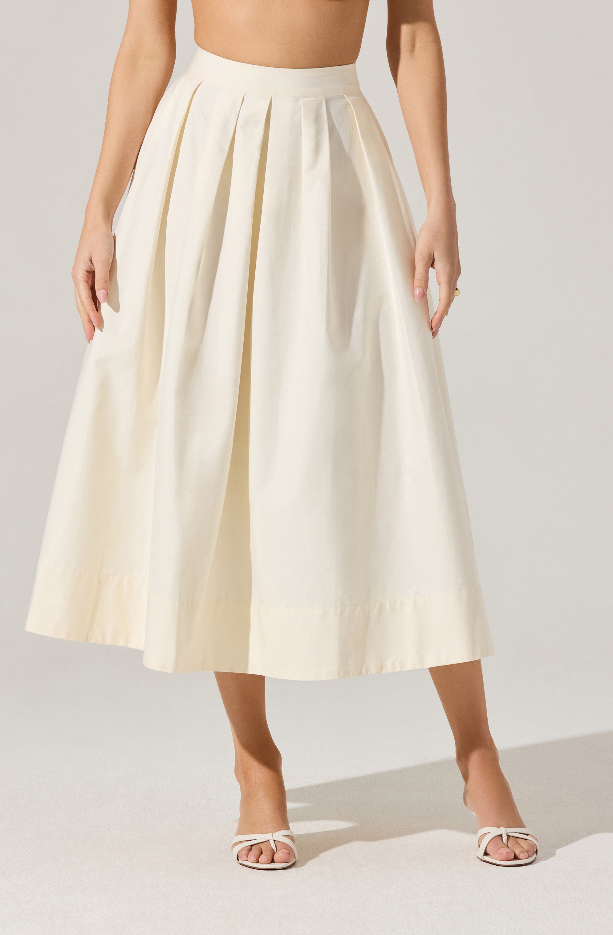 A woman is wearing a high-waisted, cream-colored, wide-leg midi skirt with pleats at the waist. The skirt has a smooth, slightly shiny texture and falls to mid-calf length. She is also wearing white open-toe heels with ankle straps.