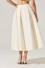 A woman is wearing a high-waisted, ankle-length white skirt with pleats running down from the waistband. The skirt is made of a smooth, slightly shiny fabric. She is also wearing a cropped yellow top and white high-heeled shoes with a pointed toe.