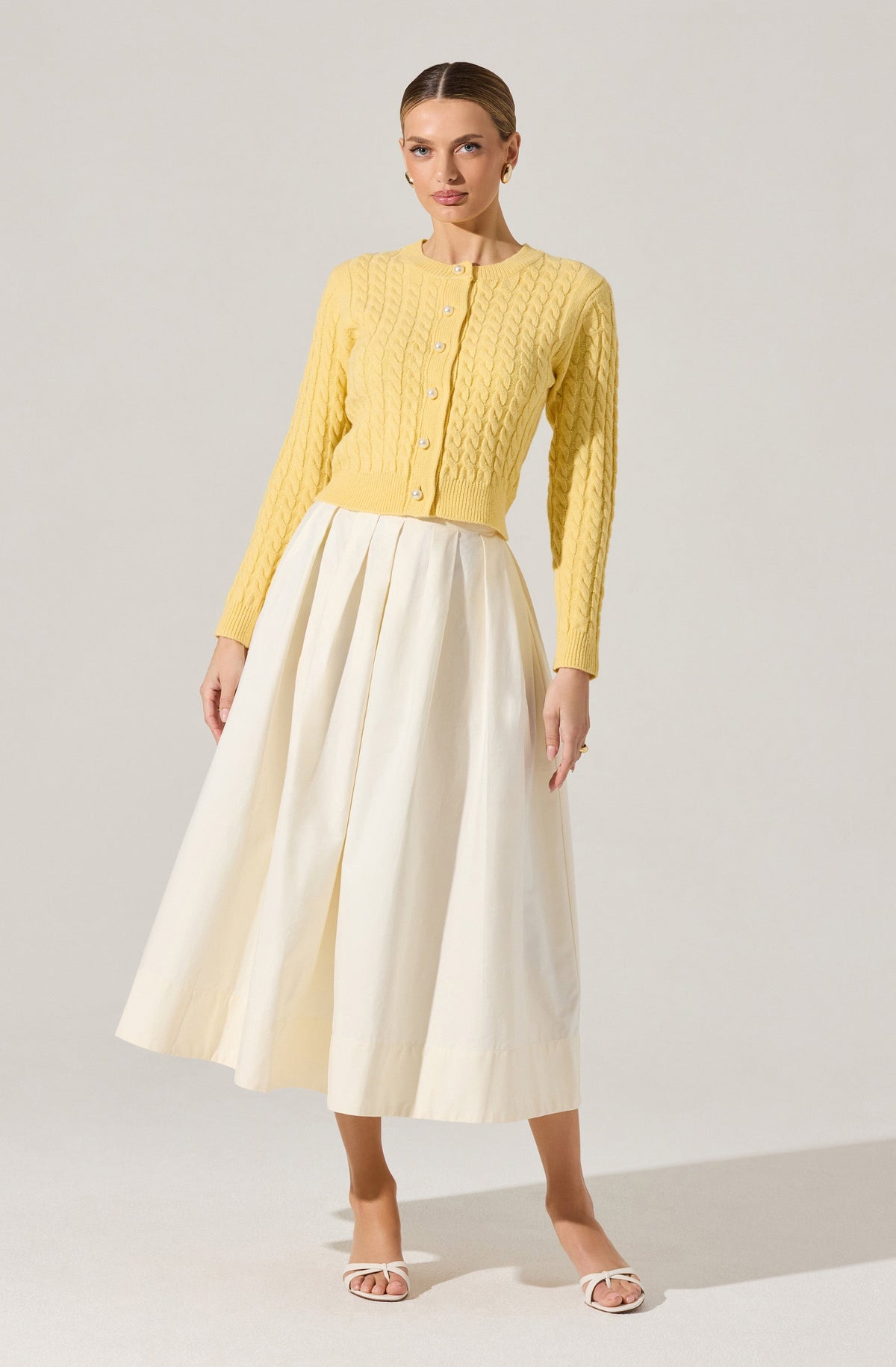 A woman is wearing a yellow, long-sleeve, button-up cardigan with a cable knit pattern. She pairs it with a high-waisted, long, cream-colored skirt that has pleats and a fluttery, flowing silhouette. She is also wearing white open-toe sandals with a small heel.