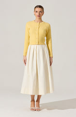 A woman is wearing a light yellow, long-sleeved, cable-knit cardigan with a round neckline and button closure down the front. She pairs it with a high-waisted, cream-colored, wide-legged midi skirt with pleats. She completes the outfit with open-toe, strappy heels.