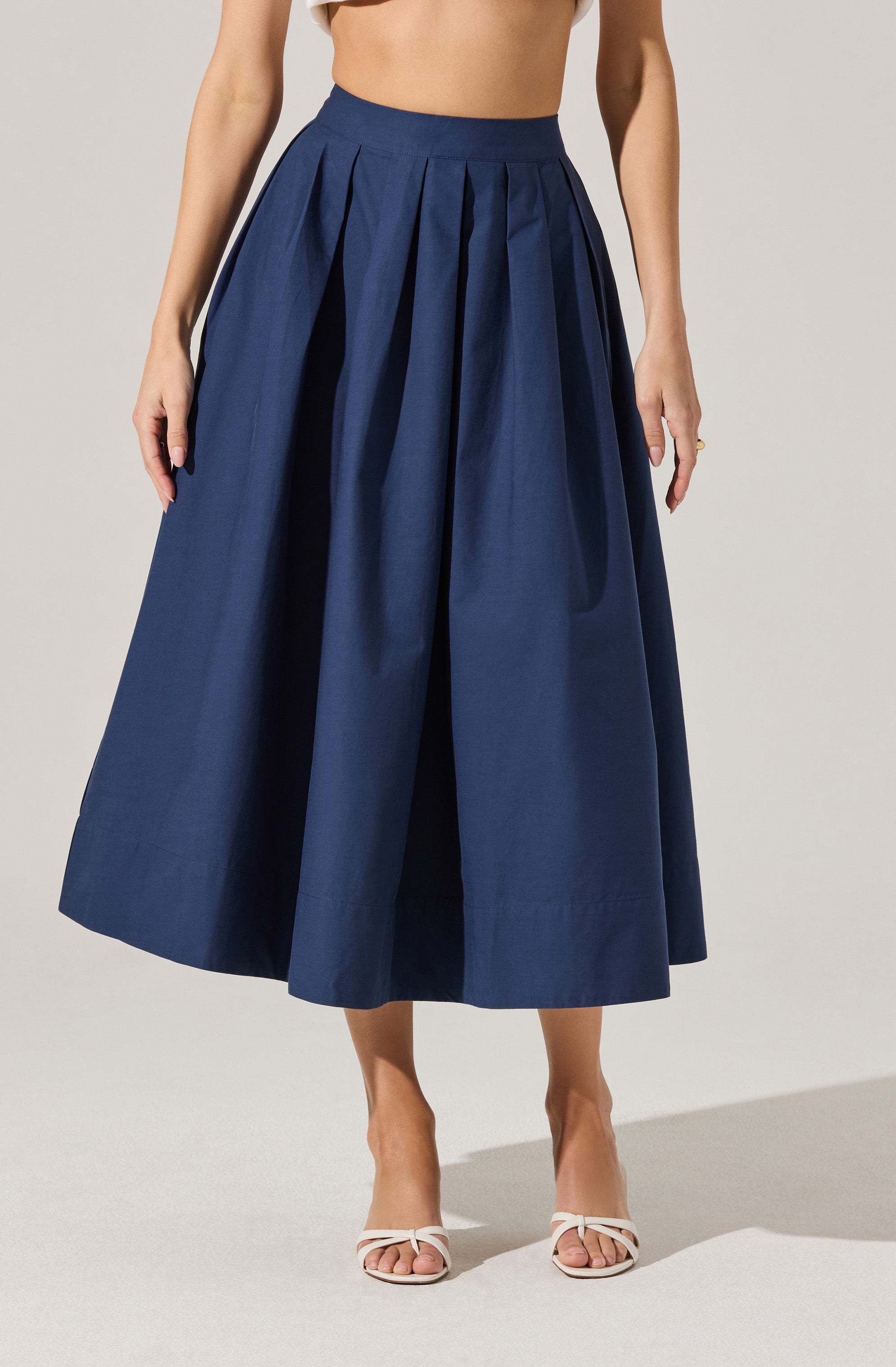 A woman is wearing a high-waisted, navy blue, full-length pleated skirt. The skirt has a wide waistband and large box pleats that add volume and structure. She pairs it with open-toed, white high-heeled sandals with thin straps. She is topless with her arms resting at her sides.