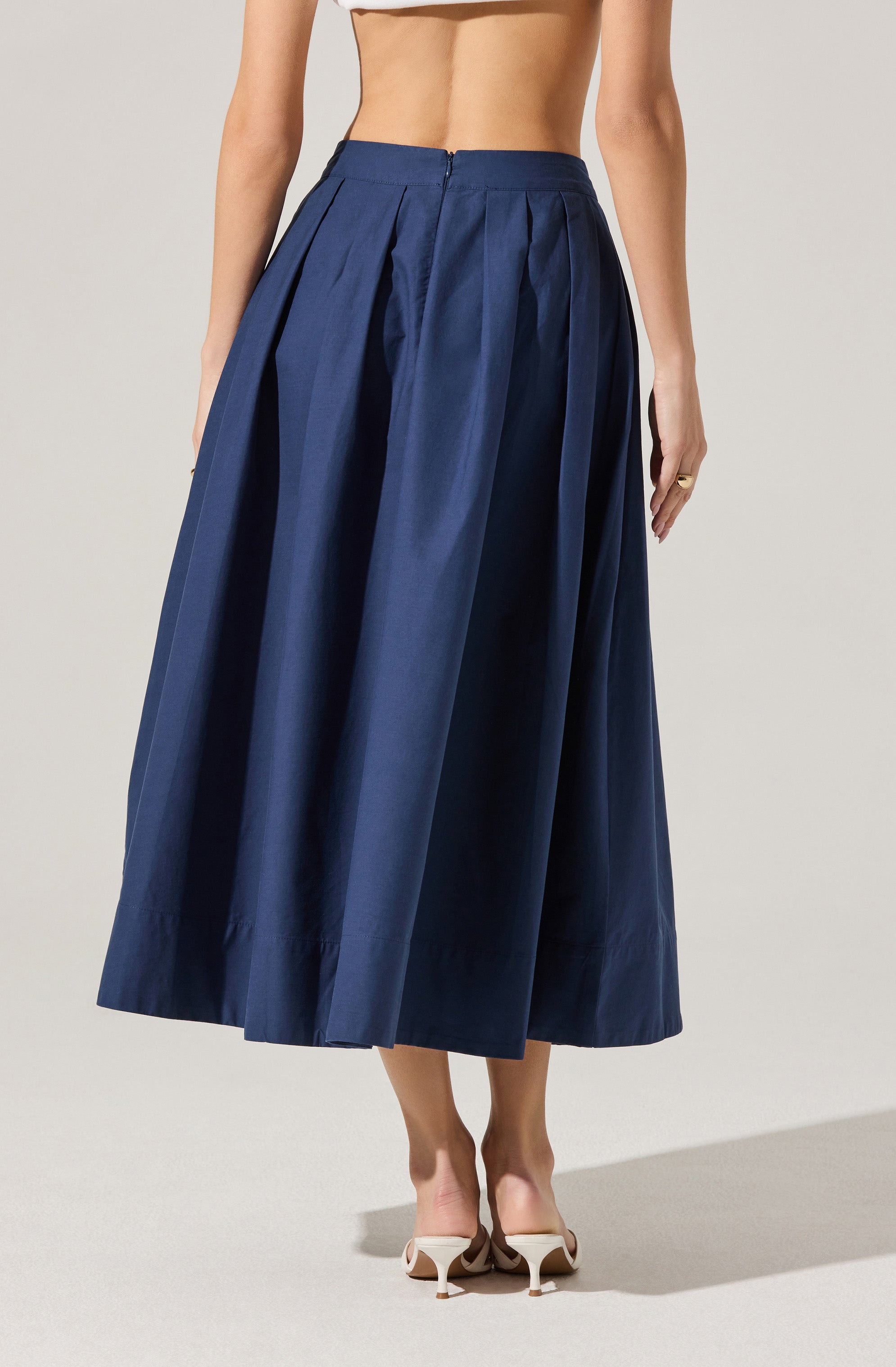 A woman is wearing a high-waisted, full-length navy blue pleated skirt made of a shiny, structured fabric. The skirt features wide pleats and a visible zipper in the back. She is also wearing beige high-heeled shoes.