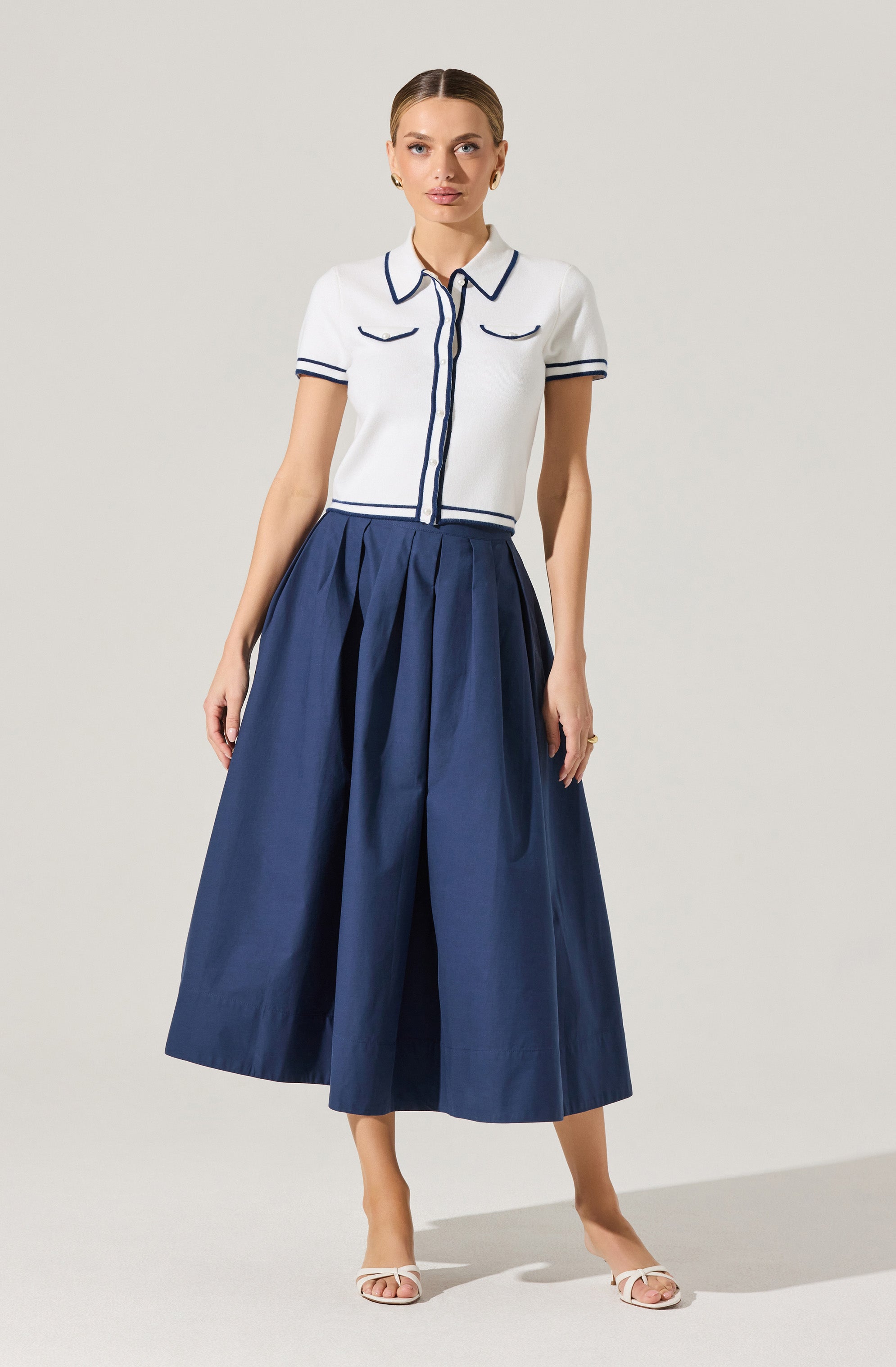 A woman is wearing a short-sleeved, collared white blouse with dark piping accents on the collar, sleeve cuffs, front pockets, and down the center. She pairs it with a high-waisted, wide-leg, pleated navy blue skirt that reaches her ankles. She completes the look with white open-toe heels.