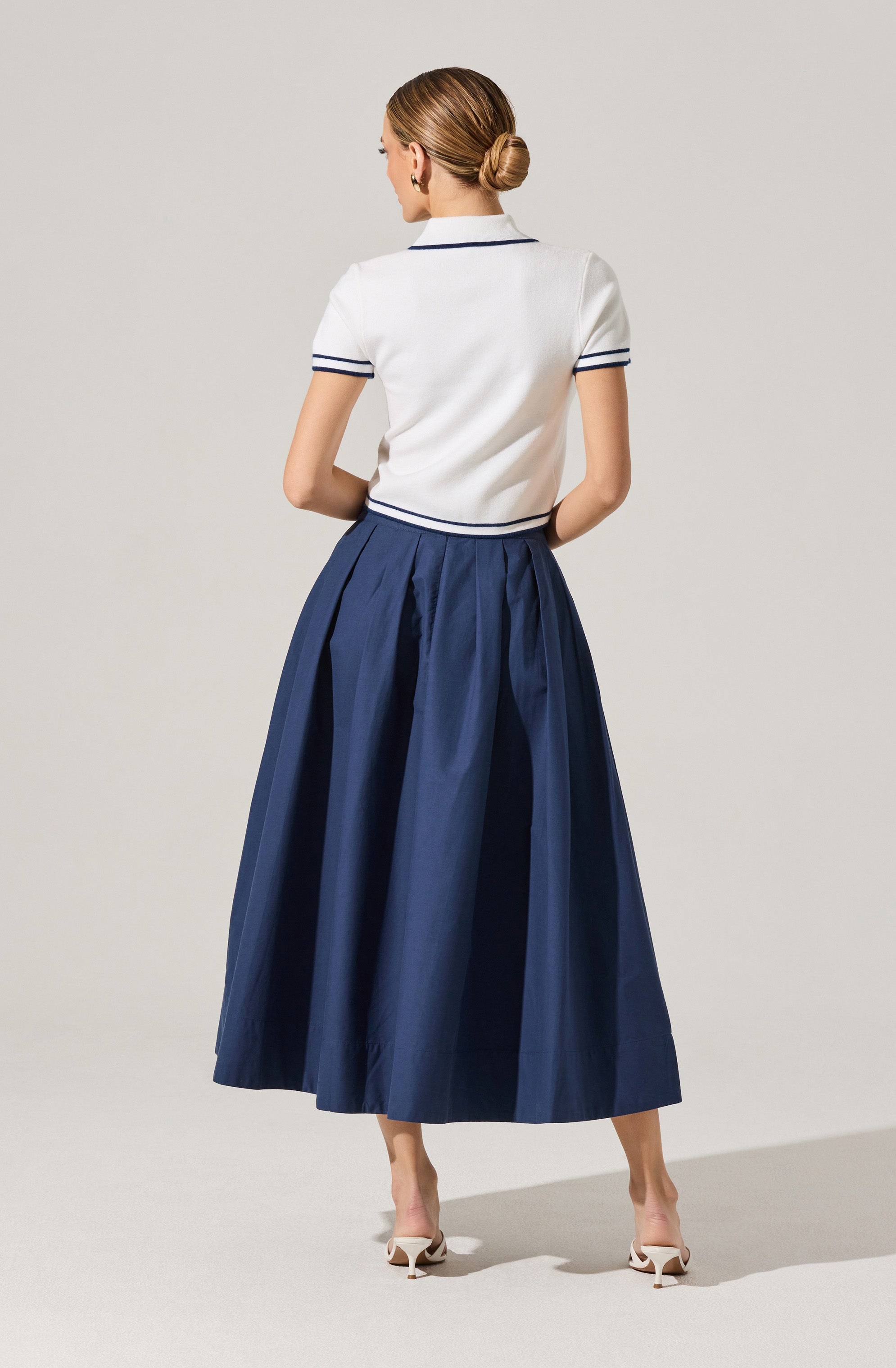 A woman is wearing a white short-sleeved top with navy blue piping on the collar and sleeve cuffs. She pairs it with a high-waisted, full-length navy blue pleated skirt. She is also wearing light-colored high-heeled shoes.