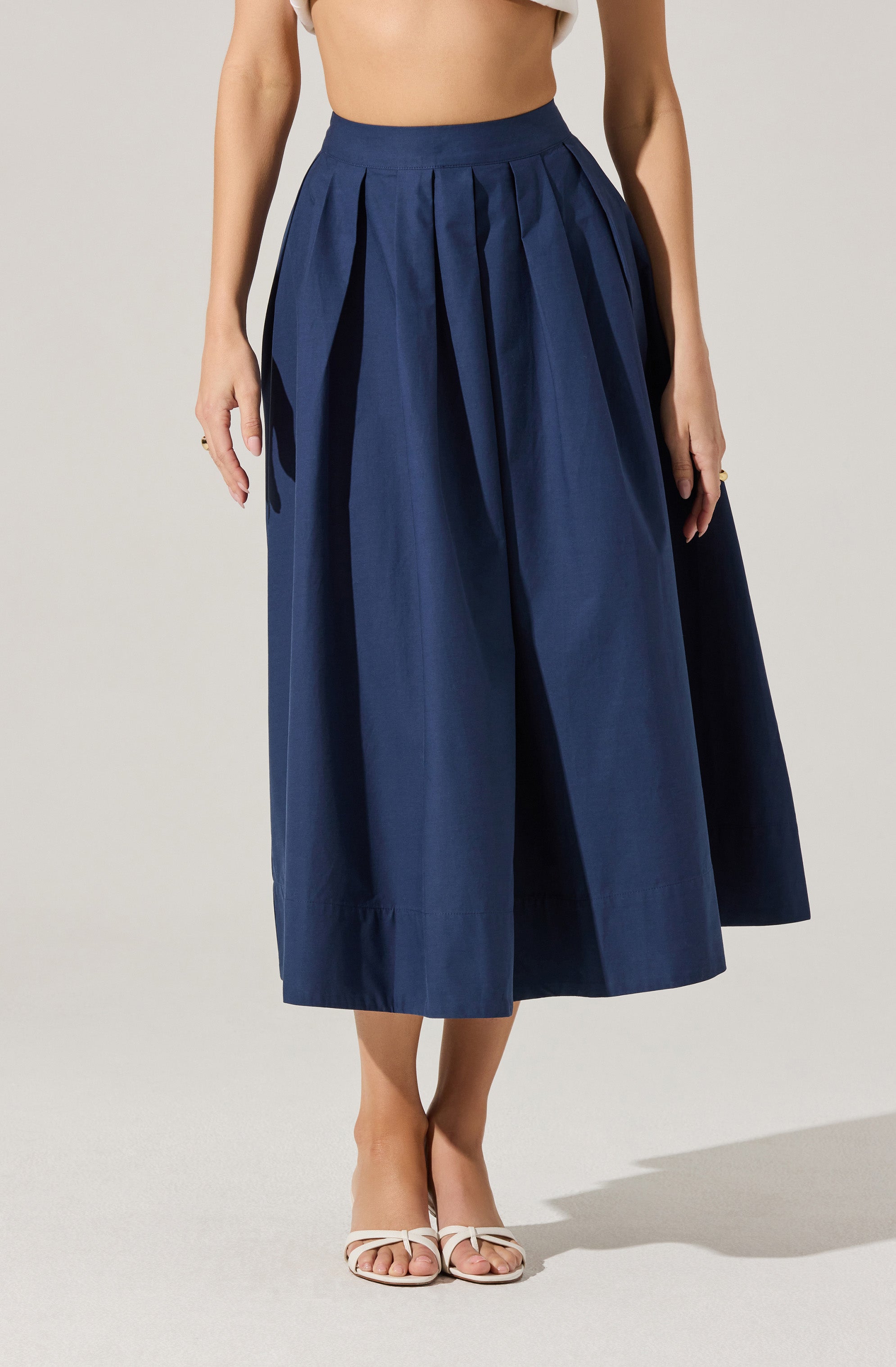 A woman is wearing a high-waisted, ankle-length navy blue skirt with wide pleats. She is also wearing white open-toe heels with ankle straps. Part of her beige-colored top is visible, but her face is not shown.
