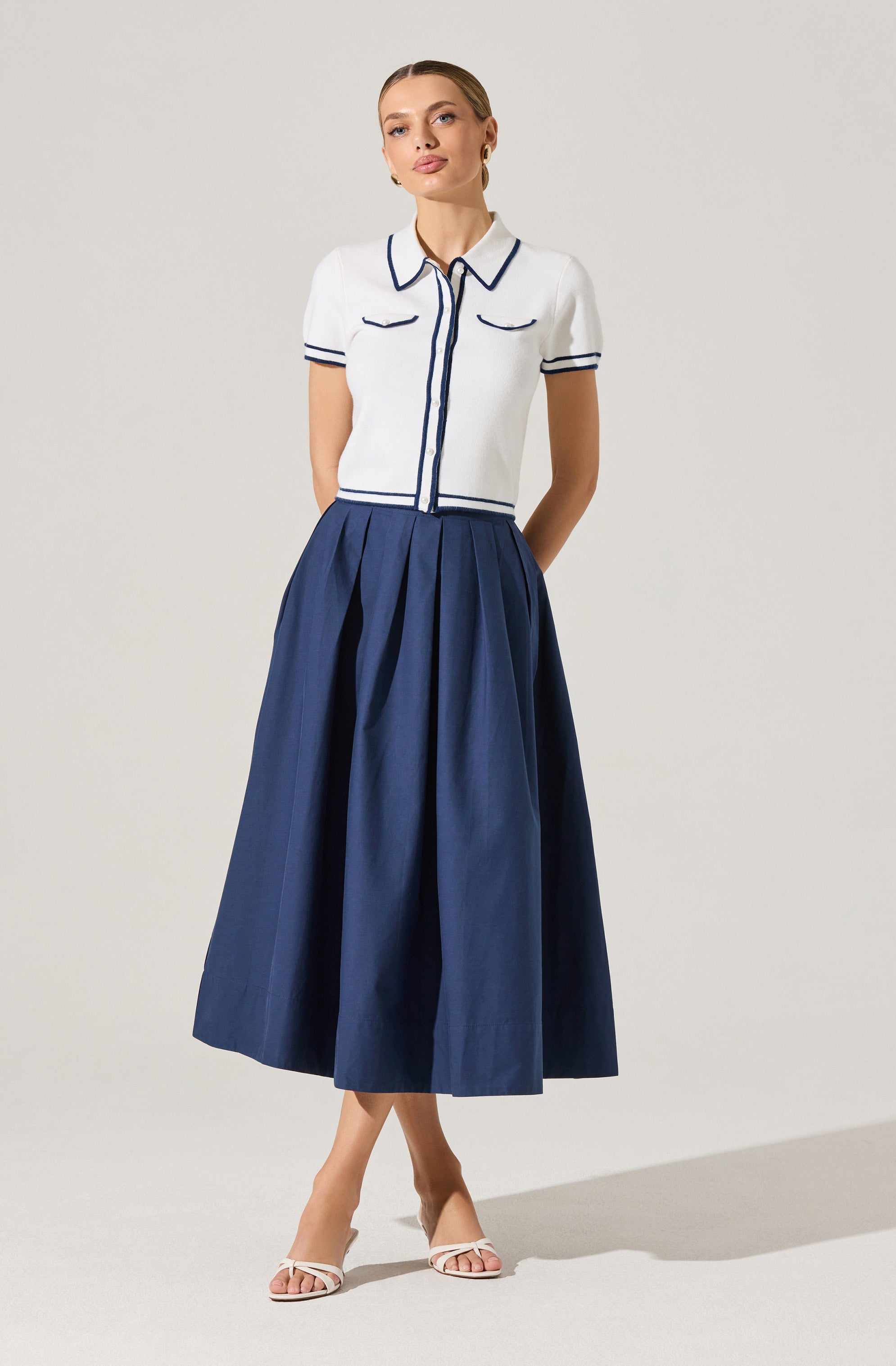 A woman is wearing a short-sleeved, white knit top with navy trim along the collar, sleeve edges, front zipper line, and pocket flaps. She pairs the top with a full, high-waisted, navy blue pleated midi skirt that falls to her ankles. She is also wearing white, open-toe, strappy high-heeled sandals.