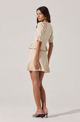 A woman is wearing a beige two-piece outfit consisting of a short-sleeved, puff-sleeve top with a peplum hem and a matching high-waisted skirt with an A-line silhouette. She is also wearing strappy high-heeled sandals.