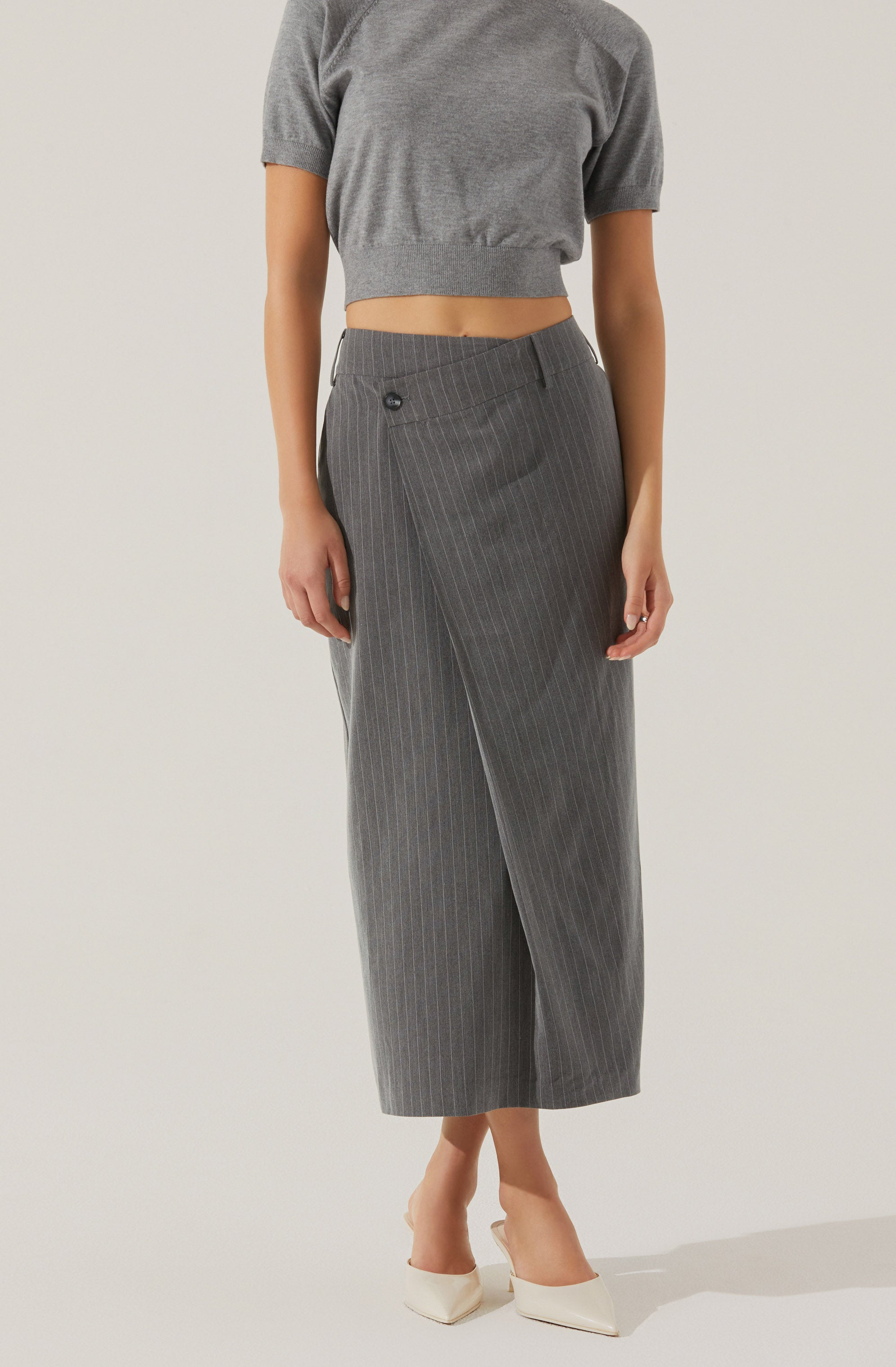 A person is wearing a loose-fitting, short-sleeved, grey crop top with a rounded neckline, paired with high-waisted, grey wide-leg trousers featuring thin vertical white pinstripes and a single button on the waistband. They are also wearing white pointed-toe high heels.