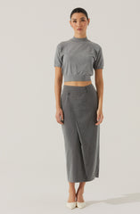 A woman is wearing a short-sleeved, gray knitted crop top with a high neckline, paired with high-waisted, gray wide-leg trousers featuring a subtle pinstripe pattern and front pleats. She is also wearing light-colored pointed-toe heels.
