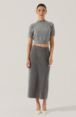A woman is wearing a grey, short-sleeved, cropped sweater with a high neckline, and high-waisted, wide-leg grey pinstripe trousers that reach her mid-calf. She is also wearing white pointed-toe shoes.