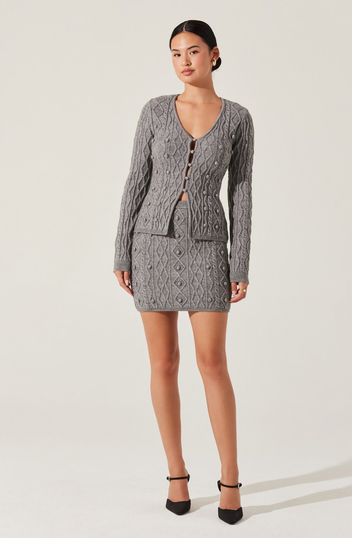 The woman is wearing a coordinated gray knit outfit consisting of a cable-knit cardigan with a V-neckline and button closure, and a matching mini skirt with a diamond and cable-knit pattern. She is also wearing black ankle-strap high heels.