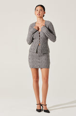 A woman is wearing a matching two-piece outfit consisting of a gray cable-knit cardigan with a front button closure and matching gray knit skirt with a diamond pattern. She is accessorizing with small hoop earrings and black high-heeled shoes with ankle straps.