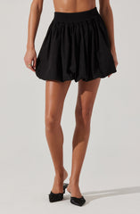 A woman is wearing a high-waisted, black, elastic waistband skirt that is voluminous and gathered, creating a bubble-like shape. The skirt is above the knees and has a loose, billowy fit. She is also wearing black flat shoes with a bow detail at the front.
