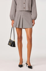 A woman is wearing a coordinated outfit consisting of a long-sleeved, cropped blazer with a small check pattern and matching high-waisted pleated mini skirt. The blazer features puffed sleeves with elastic cuffs, and the skirt has pleats at the front. She is accessorizing with a black handbag that has a gold clasp and a narrow strap. She is also wearing black high-heeled shoes with bows on the front.