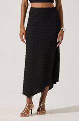 A woman is wearing a black textured two-piece outfit consisting of a sleeveless crop top and a high-waisted, asymmetrical midi skirt with a slit on one side. She has jewelry on her left hand and is wearing strappy, nude high-heeled sandals.