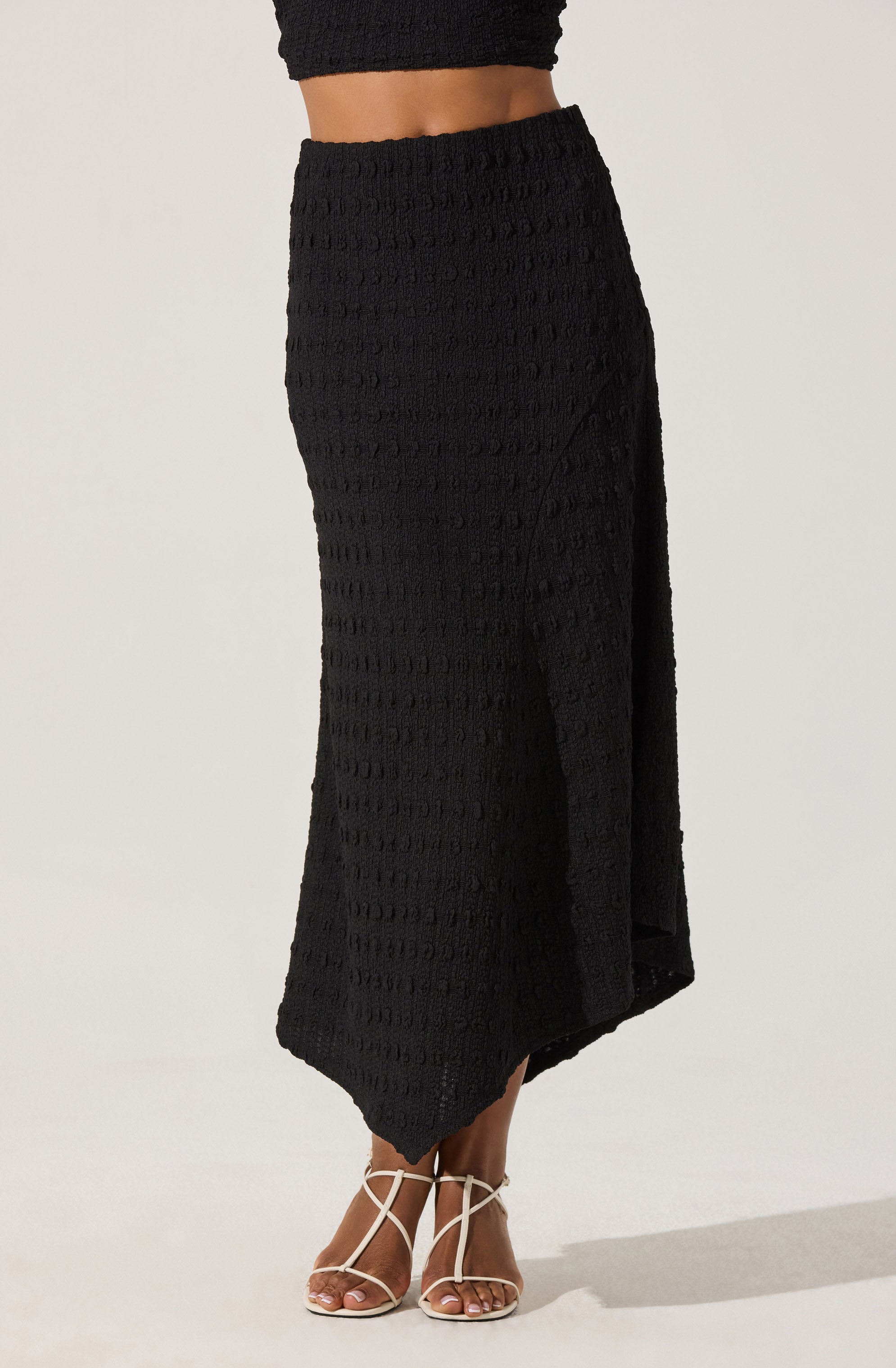 A person is wearing a black textured skirt that reaches mid-calf length, with a slit on the side. The skirt has a subtle pattern made of small raised horizontal lines. They are also wearing a matching black top that exposes the midriff. The outfit is completed with white high-heeled sandals featuring thin straps.