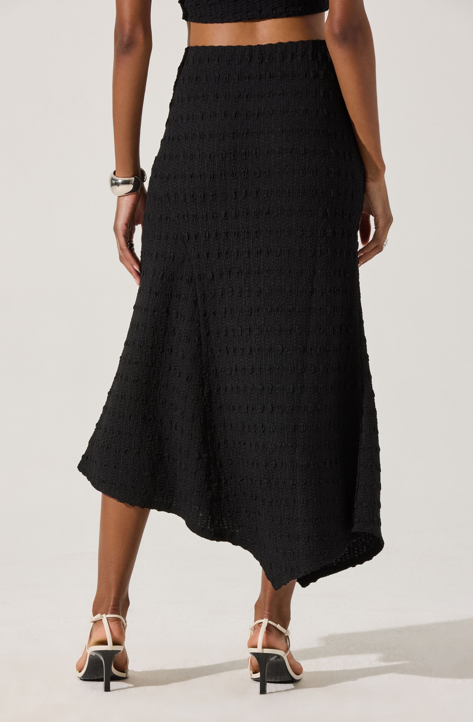 A woman is wearing a high-waisted, black, textured crochet skirt that falls to mid-calf length with an asymmetrical hemline. She has paired it with a black top and white heels with black accents. She is accessorized with a chunky silver bracelet on her left wrist.