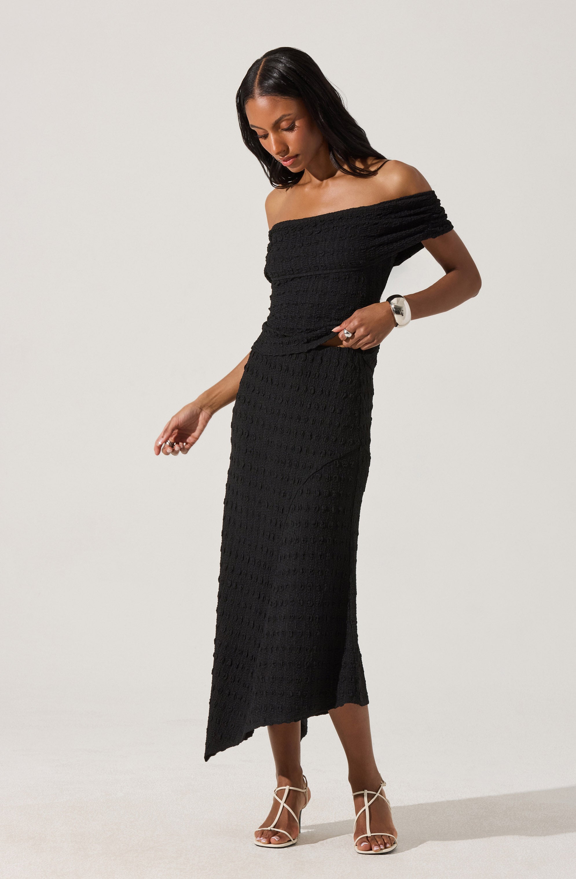 A woman is wearing a black off-the-shoulder dress made of textured fabric. The dress has a fitted bodice and a long skirt that reaches her ankles with a slit on one side. She is also wearing beige high-heeled sandals and a white wristwatch.