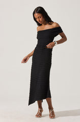 A woman is wearing a black off-the-shoulder dress made of textured fabric. The dress has a fitted bodice and a long skirt that reaches her ankles with a slit on one side. She is also wearing beige high-heeled sandals and a white wristwatch.
