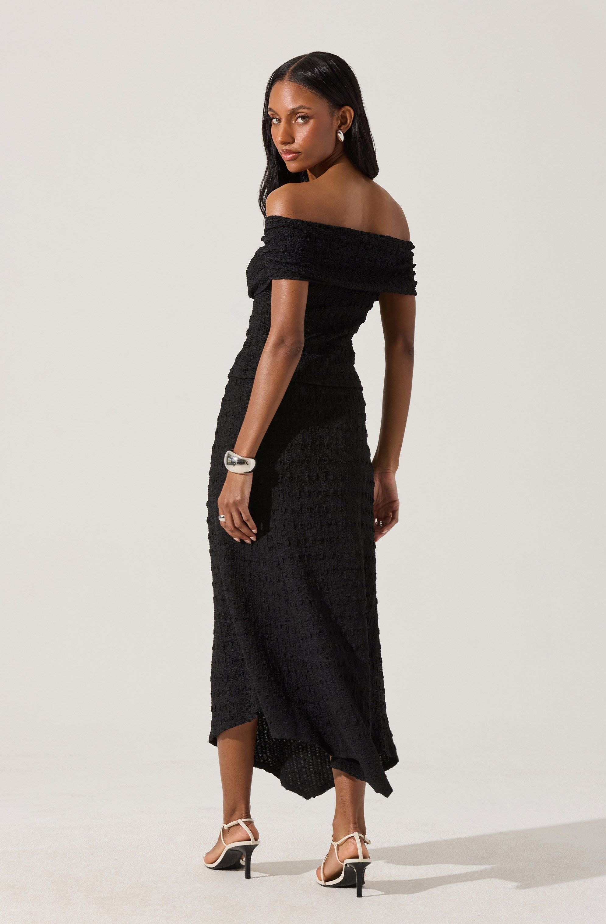 A woman is wearing an off-the-shoulder black crocheted dress that is fitted through the waist and hips, with a slightly flared hem that falls to her ankles. She has paired the dress with white high-heeled sandals with thin ankle straps and a chunky white bracelet on her wrist.