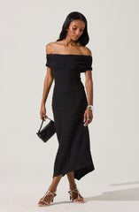 A woman is wearing a black off-the-shoulder, knitted dress that fits snugly to her body and extends down to her ankles. She accessorizes with a silver bracelet, rings, a small black handbag, and strappy high-heeled sandals.