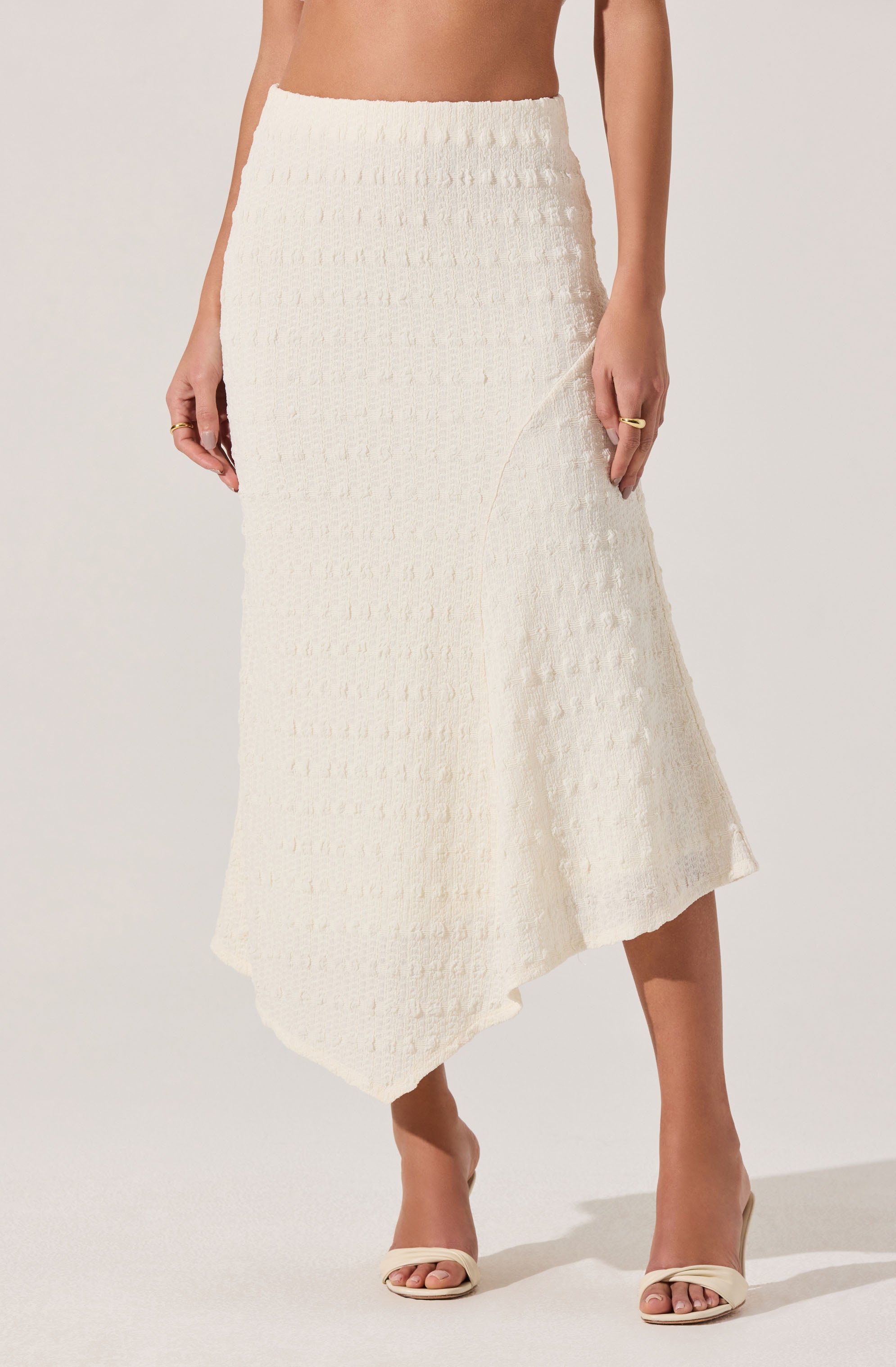 A woman is wearing a high-waisted, white textured knit skirt that features a subtle geometric pattern and an asymmetrical hemline. She has paired it with open-toe, cream-colored heels and gold rings on her fingers.
