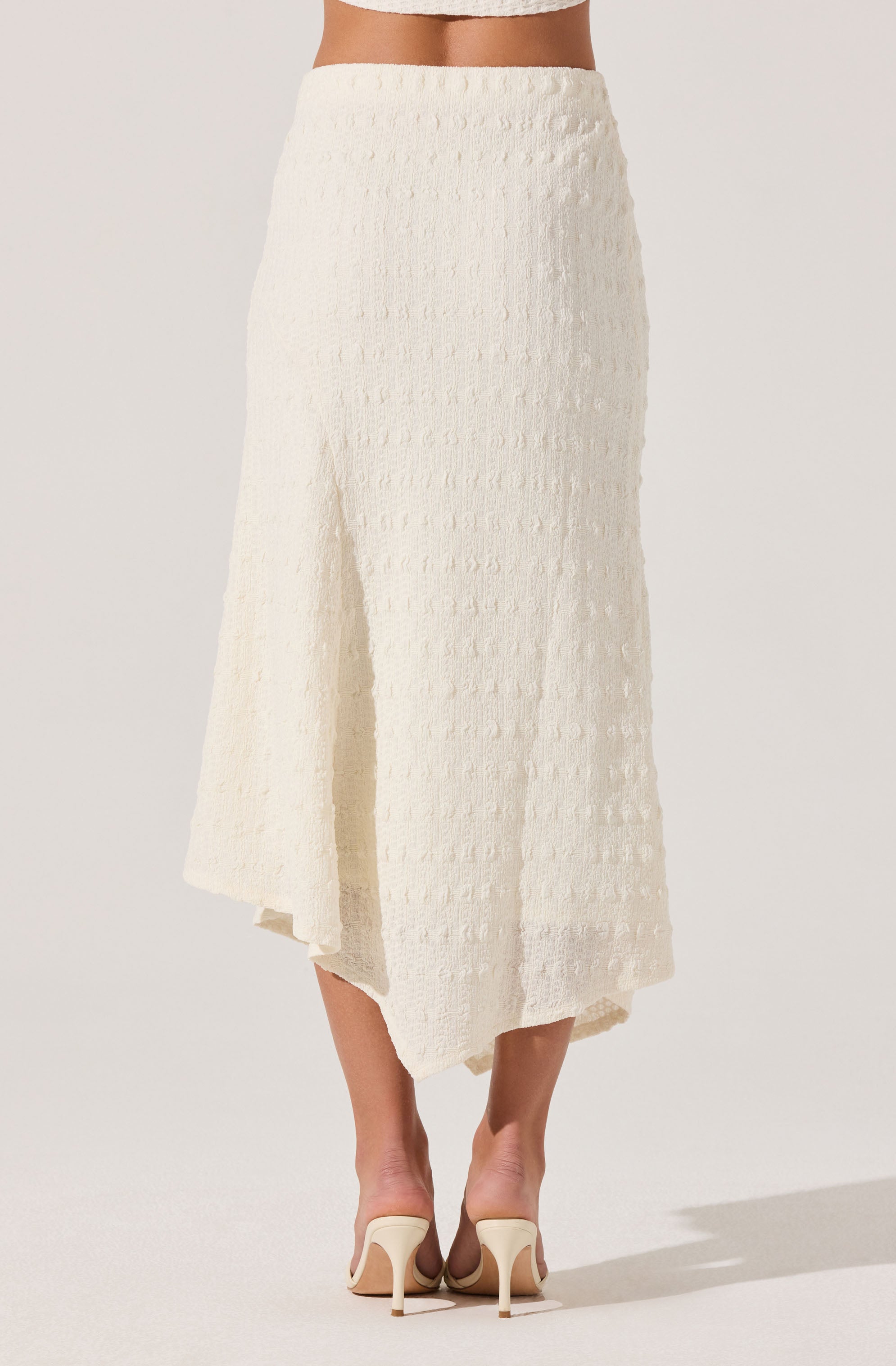 A woman is wearing a high-waisted, ankle-length cream-colored skirt with a textured, dotted pattern. The skirt features a slit on the side, revealing part of her leg. She pairs it with matching cream-colored high-heeled shoes.