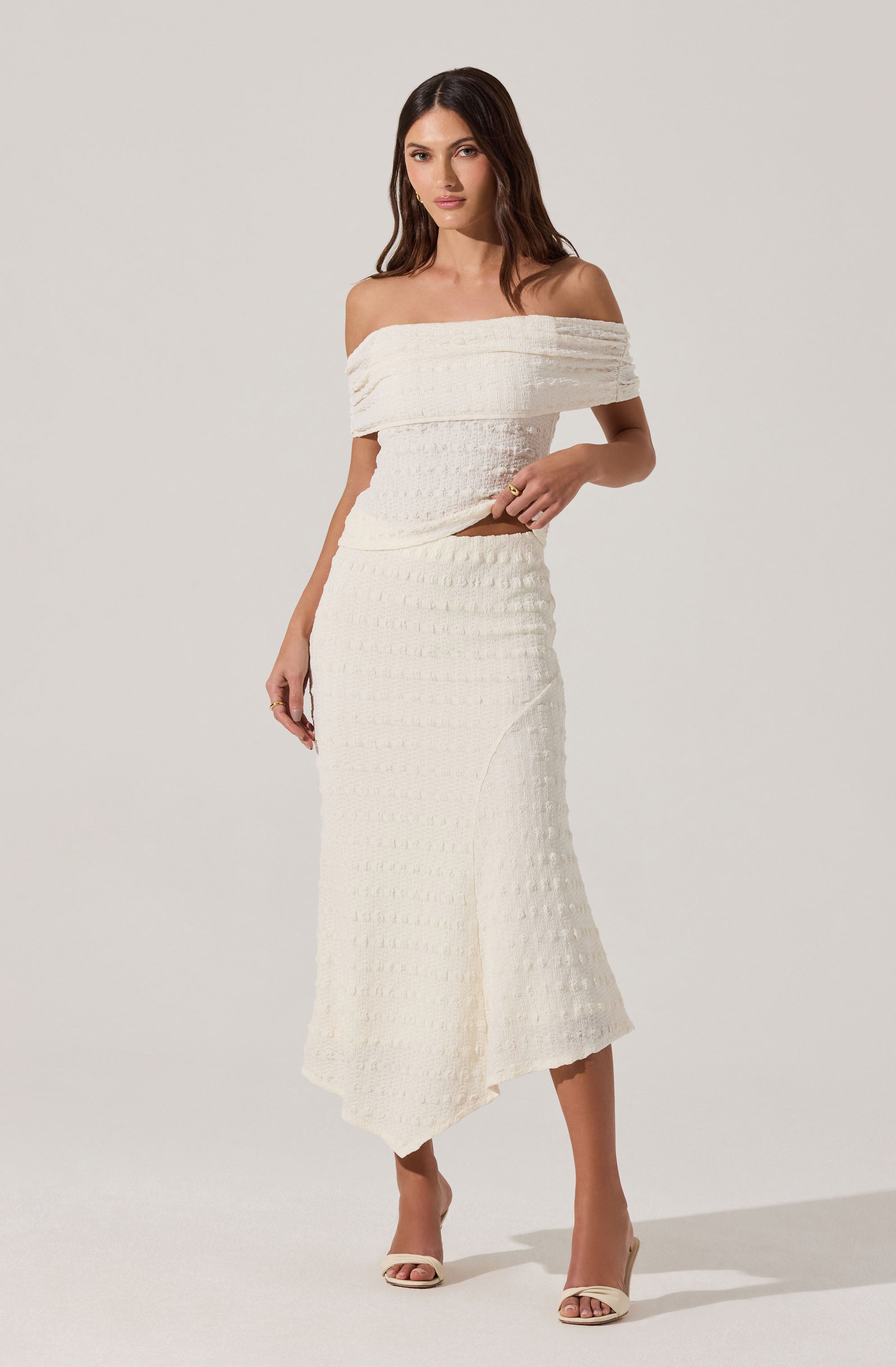 A woman is wearing an off-the-shoulder, cream-colored knit top with horizontal ridges and a matching high-waisted, ankle-length skirt featuring a textured pattern and a side slit. She is also wearing cream-colored open-toe heels.