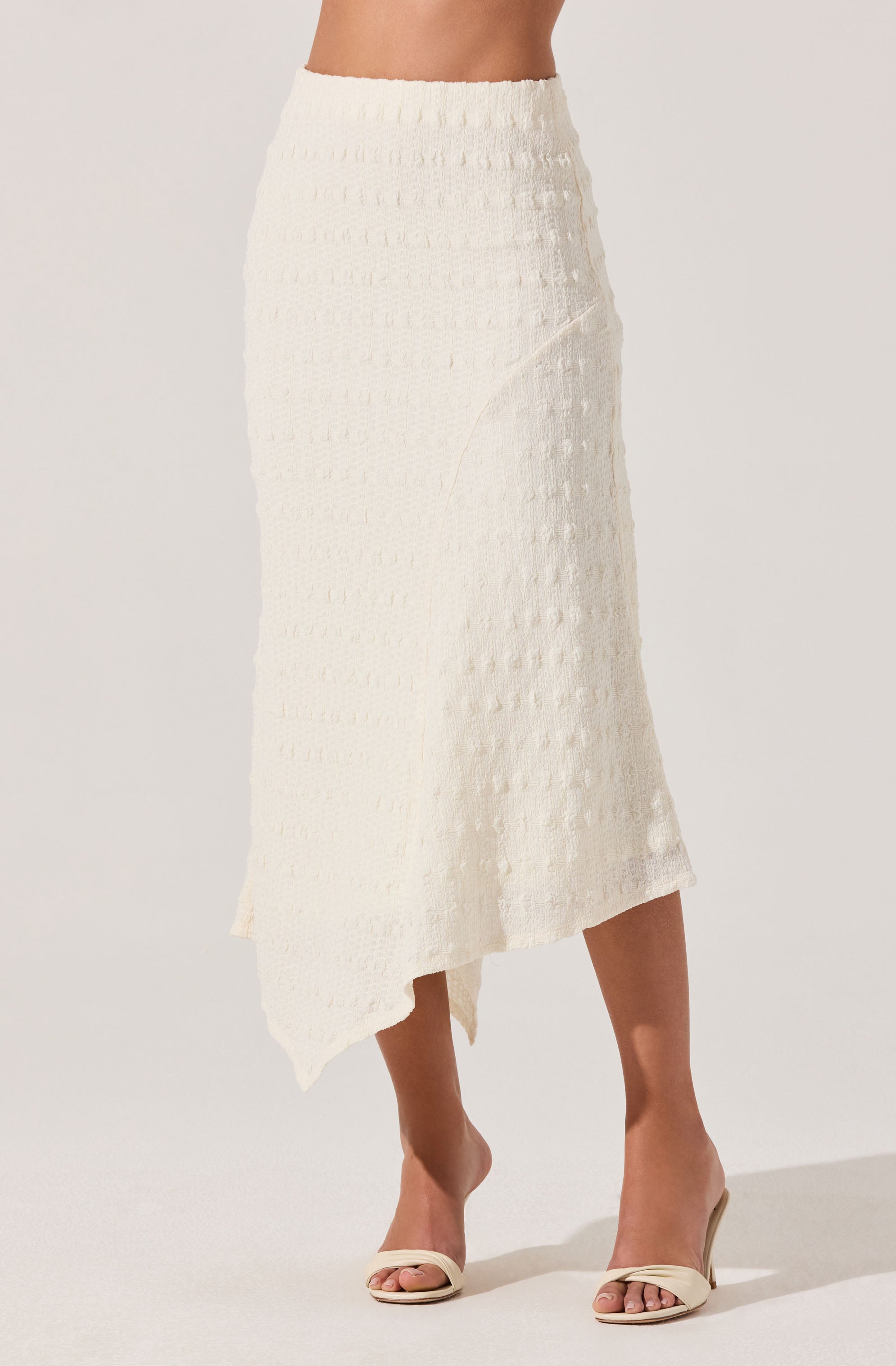 A woman is wearing a cream-colored, textured, midi-length skirt with an asymmetrical hemline. She pairs it with open-toe, ankle-strap high heels in a matching cream color. The skirt features a pattern of small, raised rectangular designs throughout.