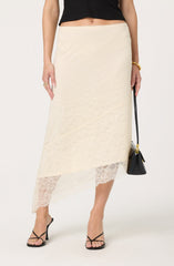 A woman is wearing a black sleeveless top paired with a cream-colored lace skirt that falls below the knees. The skirt features a sheer lace hemline with intricate floral patterns. She accessorizes with a gold bangle, multiple rings, and is holding a black handbag with a gold clasp. She is also wearing black high-heeled sandals with thin straps.