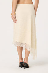 A woman is wearing a cream-colored, asymmetrical lace skirt that falls to mid-calf length. The skirt features a delicate lace overlay with a floral pattern and scalloped lace trim at the hem. She is also wearing black high-heeled sandals with an open toe and ankle strap, and a gold bracelet on her right wrist.