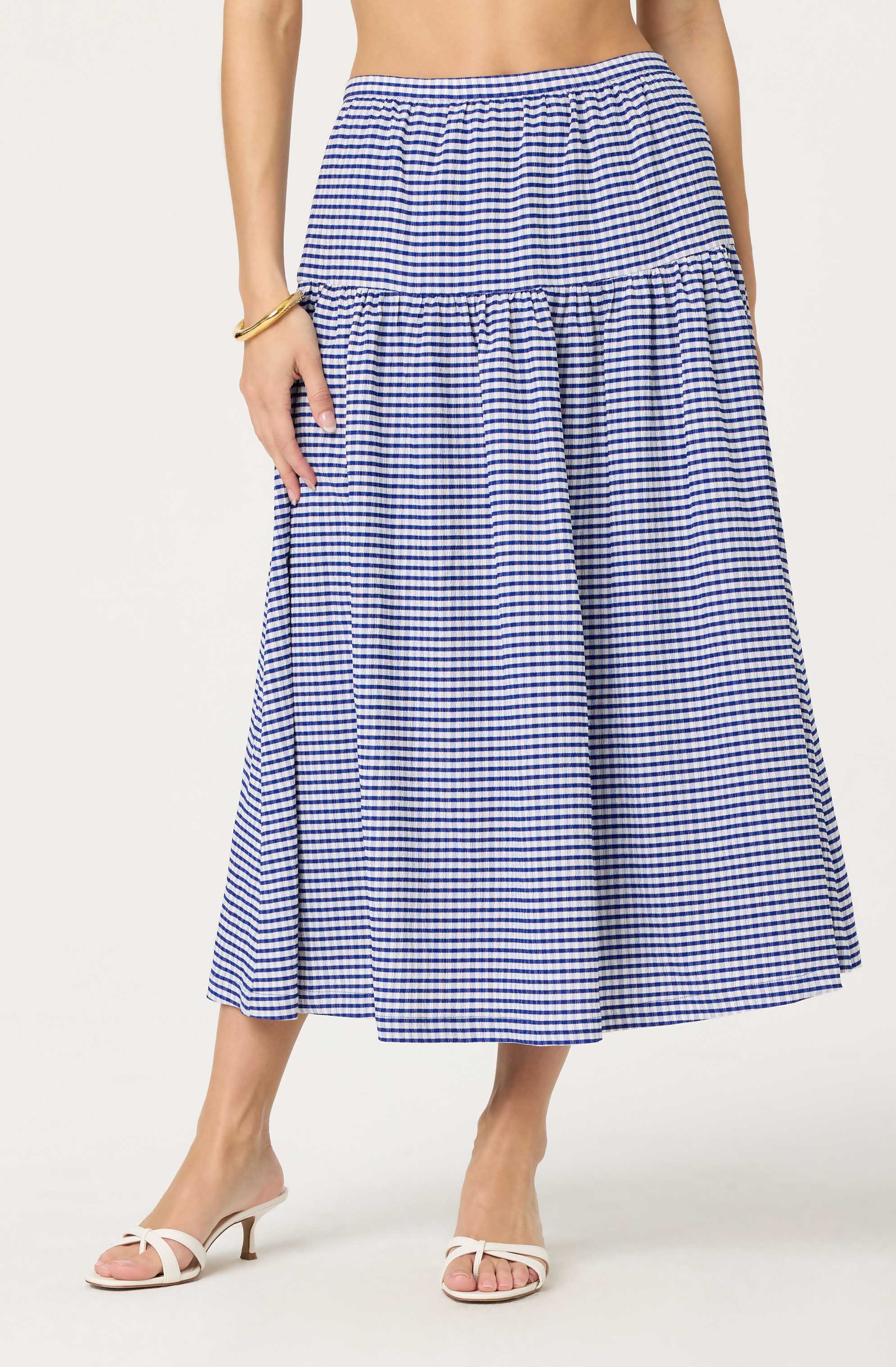 A woman is wearing a blue and white gingham checkered maxi skirt with a high waist. The skirt has a tiered design, with a gathered section at the top and a wider, flowing section below. She is also wearing white high-heeled sandals with a bow detail on the front. She has a gold bracelet on her left wrist.