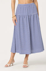 A woman is wearing a blue and white gingham checkered maxi skirt with a high waist. The skirt has a tiered design, with a gathered section at the top and a wider, flowing section below. She is also wearing white high-heeled sandals with a bow detail on the front. She has a gold bracelet on her left wrist.