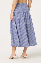 A woman is wearing a long, high-waisted skirt with a blue and white gingham check pattern. The skirt has a gathered, tiered design that creates a flowing silhouette. She is also wearing white high-heeled sandals with a thin strap across the toes and a small heel.