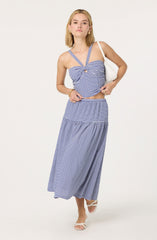 A woman is wearing a two-piece blue and white gingham outfit consisting of a fitted, sweetheart neckline crop top with wide straps and a cut-out detail in the center, paired with a high-waisted, ankle-length skirt with an elastic waistband and a ruffled, tiered design. She is accessorizing with gold earrings, a gold bracelet on her right wrist, and is carrying a white shoulder bag. She is also wearing white open-toe sandals with a low heel.