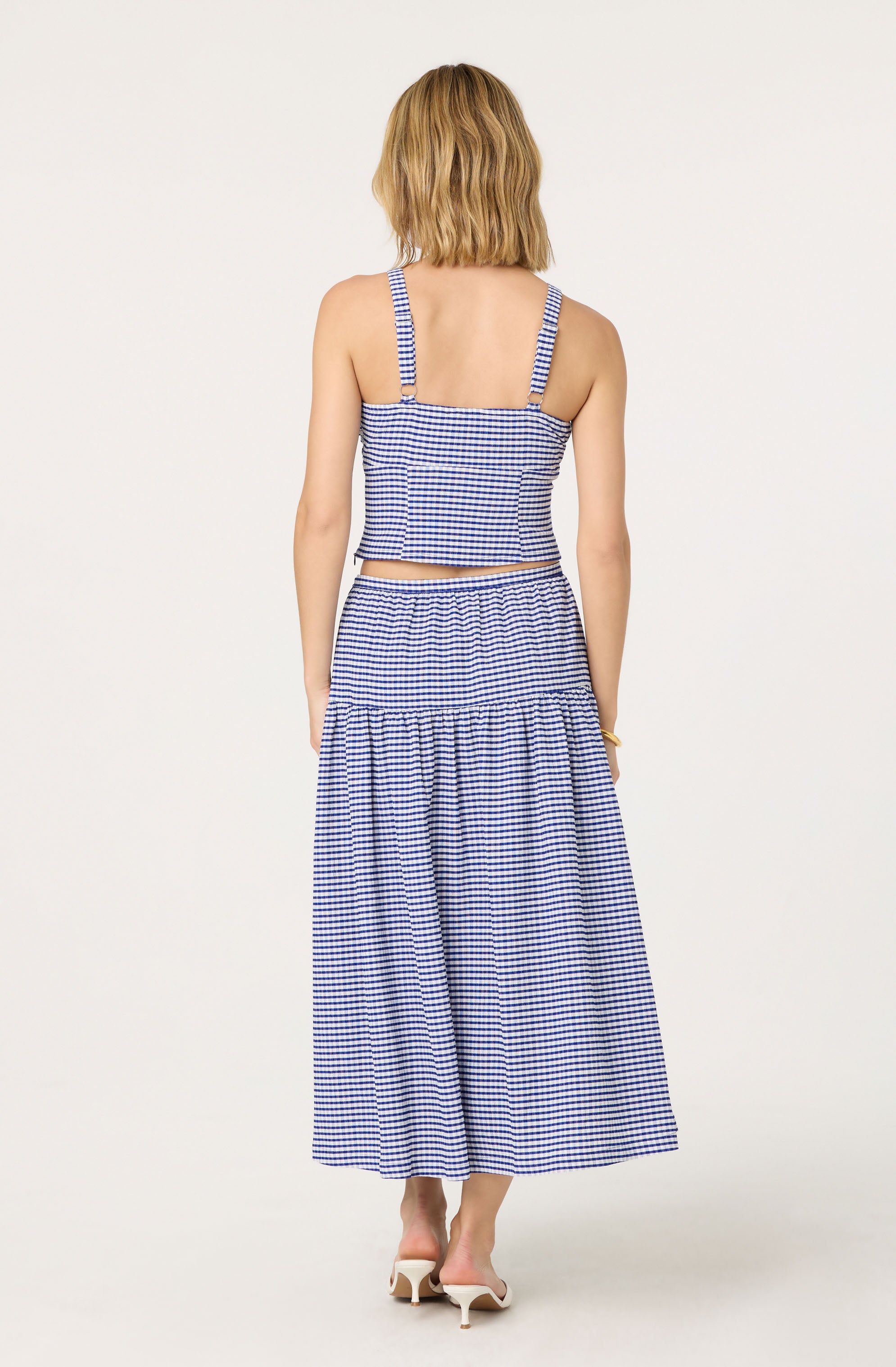 A woman is wearing a two-piece outfit made of blue and white gingham fabric. The top is a sleeveless, fitted crop top with adjustable straps. The matching skirt is a high-waisted, full-length style with a gathered section near the hem, creating a tiered effect. She is also wearing white heeled shoes.