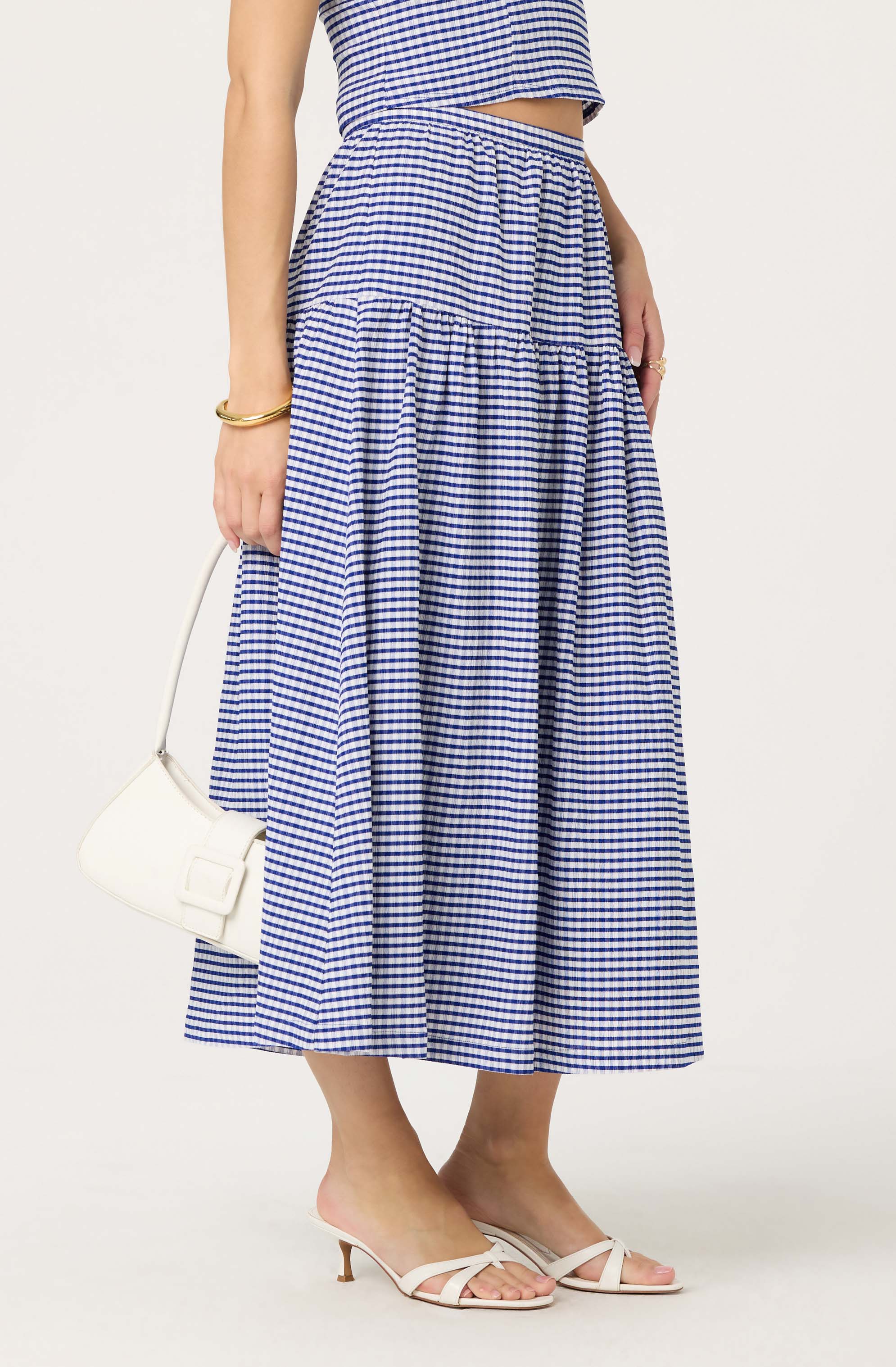 The image shows a woman wearing a two-piece outfit with a blue and white checked pattern. The top is a sleeveless crop top with a straight hem, and the skirt is a high-waisted, ankle-length, gathered skirt with the same pattern. She accessorizes with a gold bracelet, rings, and carries a small white handbag. She is also wearing white strappy heeled sandals.