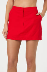 A person is wearing a bright red, high-waisted skirt that is short in length. The skirt has a smooth, fitted appearance and features a hidden front zipper. The person has one hand in their pocket and the other hanging by their side.