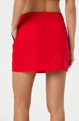 A person is wearing a bright red mini skirt with a smooth, fitted waistband. The skirt is made of a solid fabric with a slight sheen and has a clean, simple design. The individual has their hands in pockets on either side of the skirt. The background is plain and light-colored.