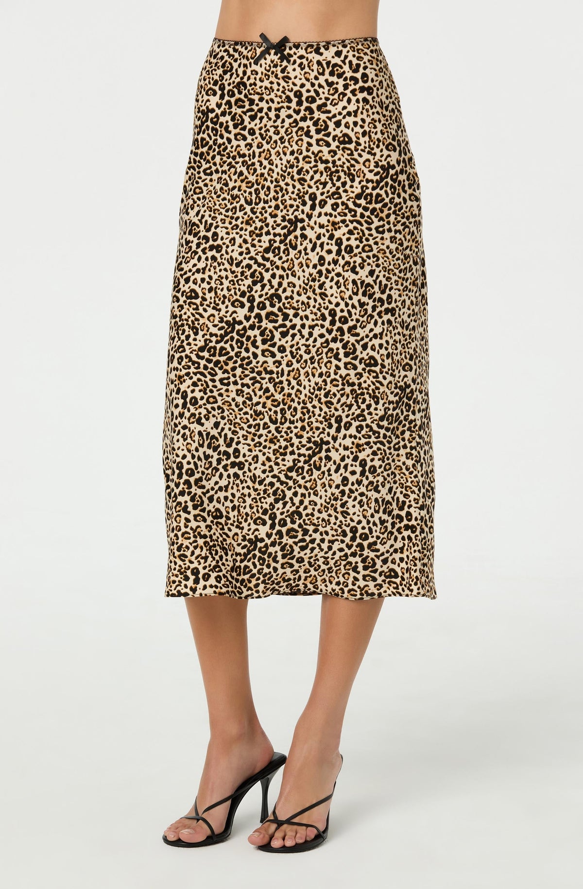 A woman is wearing a long, high-waisted skirt with a leopard print pattern in shades of tan and black, featuring a small black bow at the waistband. She pairs the skirt with black high-heeled sandals.