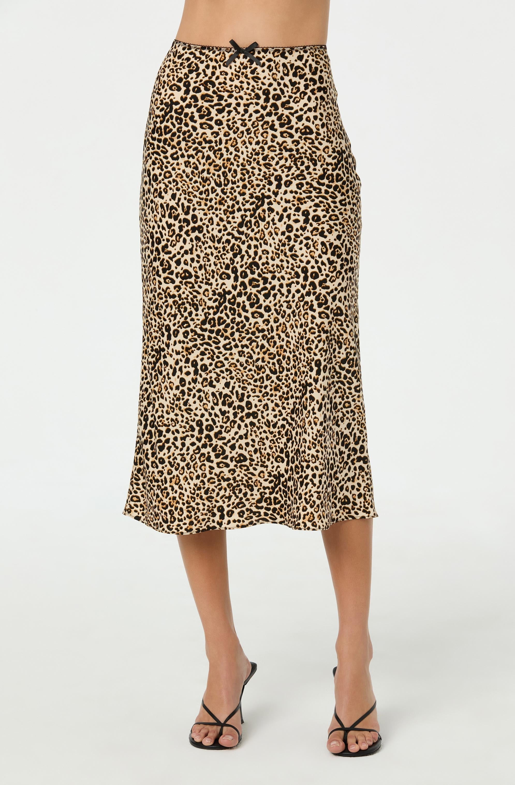 A woman is wearing a midi-length skirt with a leopard print pattern in shades of tan, black, and beige. The skirt has a fitted waist with a small black bow at the center front. She is also wearing black strappy high-heeled sandals.
