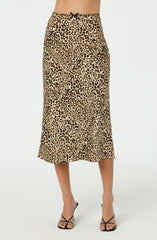 A woman is wearing a midi-length skirt with a leopard print pattern in shades of tan, black, and beige. The skirt has a fitted waist with a small black bow at the center front. She is also wearing black strappy high-heeled sandals.