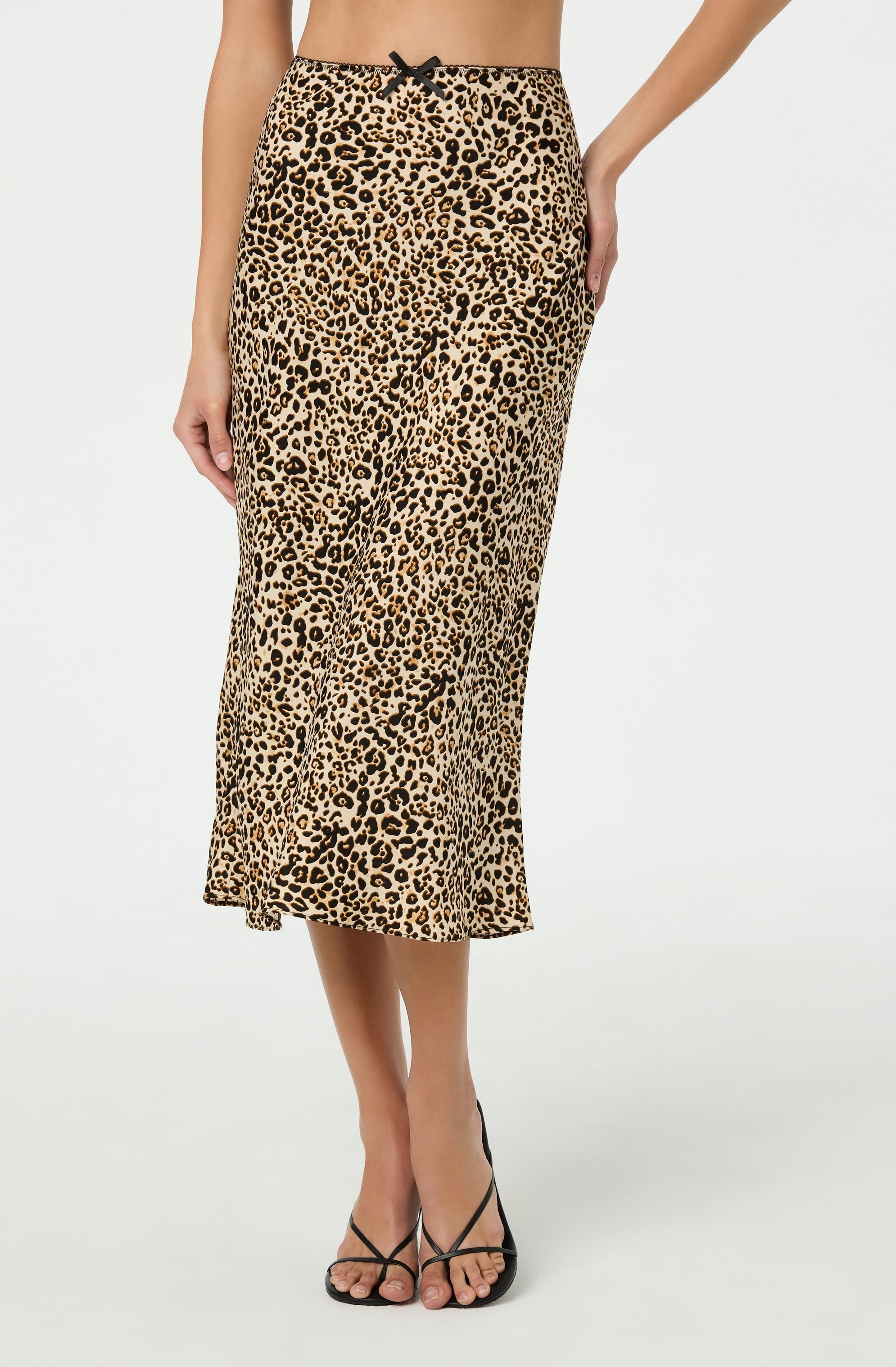 A woman is wearing a midi-length skirt with a leopard print pattern featuring black spots on a tan background. The skirt has a black bow at the waist and appears to be made of a lightweight, flowy fabric. She is also wearing black strappy high-heeled sandals.
