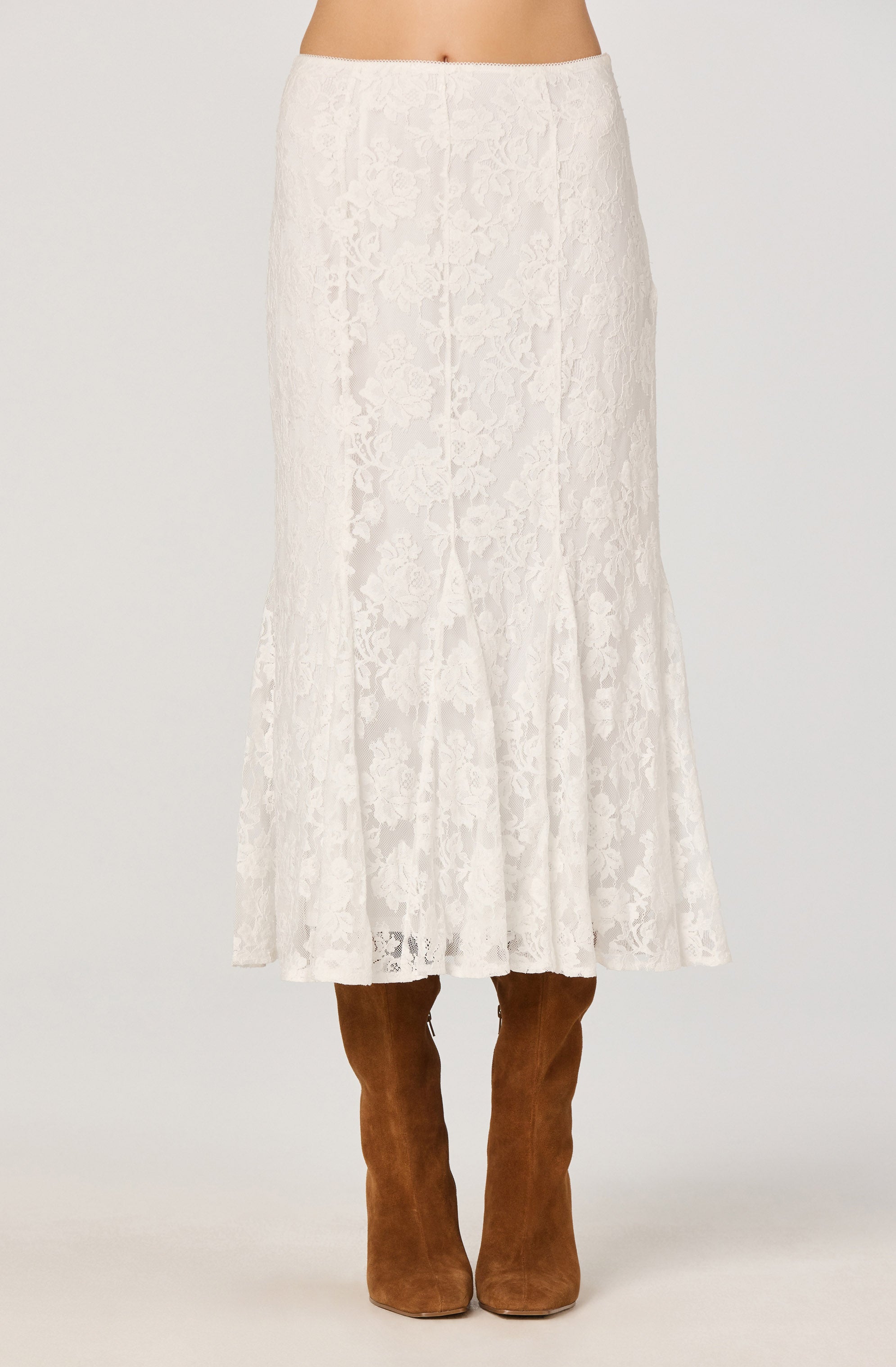 A woman is wearing a long, white lace skirt with a floral pattern that reaches down to her ankles. She pairs the skirt with knee-high brown suede boots. The waistband of the skirt sits at her natural waist.