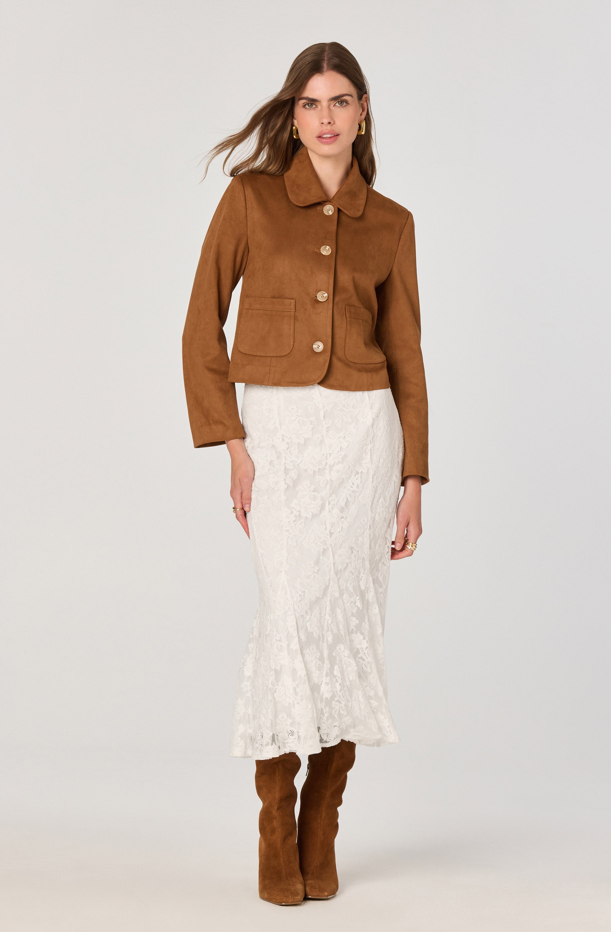 A woman is wearing a brown, long-sleeved, button-up jacket with a collar and front pockets. She pairs the jacket with a long, white, textured skirt that reaches her ankles, and matching brown knee-high boots.