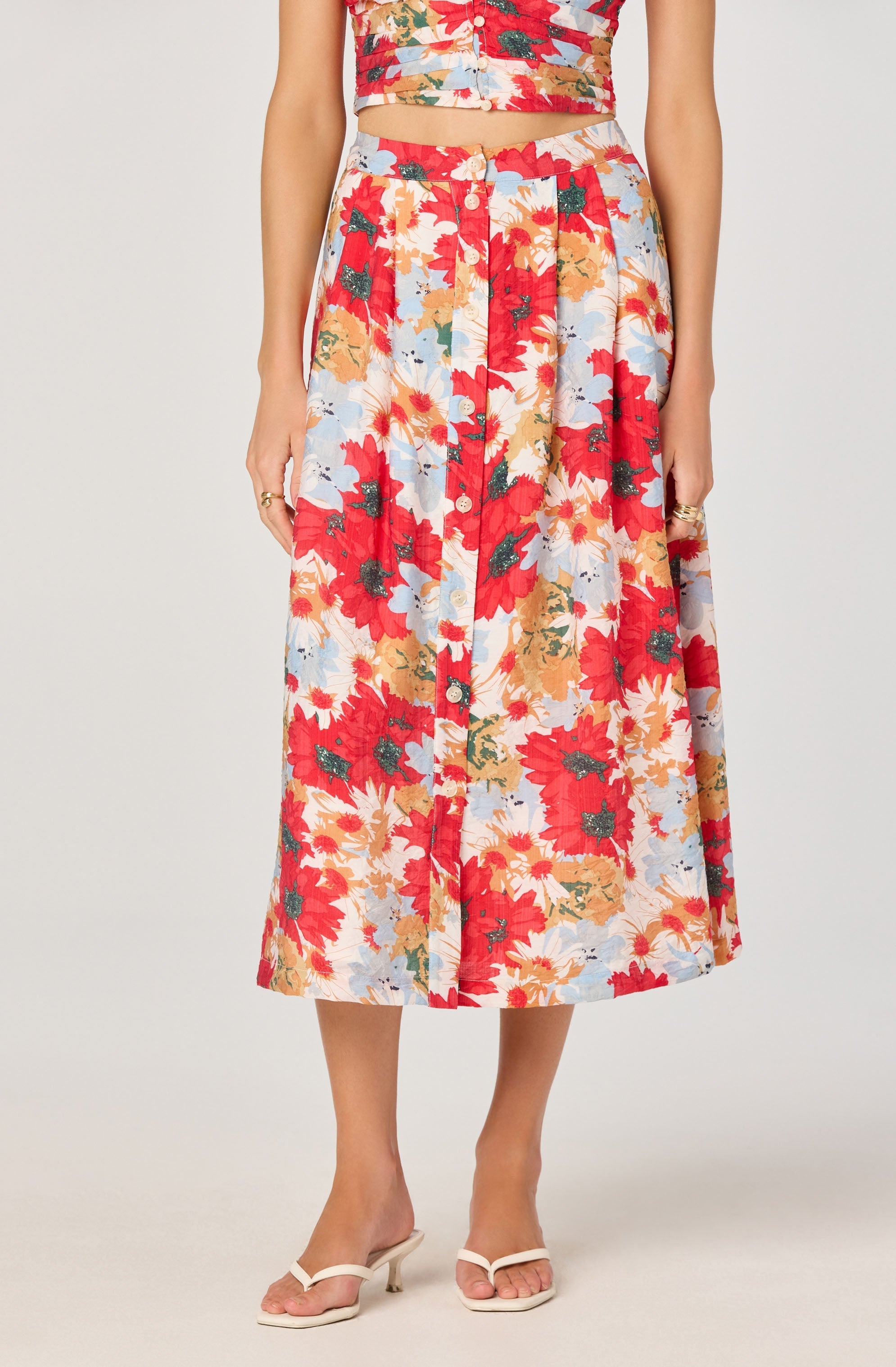A woman is wearing a matching two-piece outfit consisting of a crop top and a midi skirt. The fabric features a colorful floral pattern with large red, orange, cream, and light blue flowers and green leaves. She is also wearing beige heeled sandals.