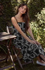 A woman is seated outdoors on a wooden chair next to a small wooden table. She is wearing a black sleeveless dress with thin spaghetti straps, featuring a white floral and geometric pattern. She has long, wavy brown hair and is accessorized with gold hoop earrings and rings.