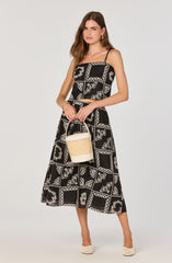 A woman is wearing a black sleeveless jumpsuit with thin spaghetti straps. The jumpsuit features a white floral and geometric pattern throughout. She is holding a beige woven handbag with a white bottom and a rounded handle. She is accessorized with gold hoop earrings and a gold bracelet. She is wearing white woven high-heeled shoes.